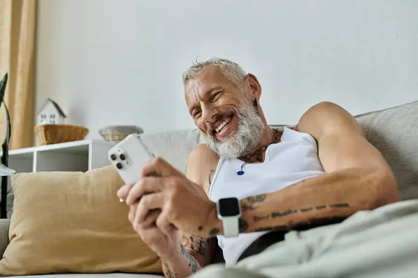 A tattooed man with a grey beard relaxes on a couch and smiles while looking at his phone. — Stock Photo