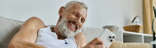 A mature gay man with tattoos and a grey beard is relaxing on a couch at home, smiling as he looks at his phone. — Stock Photo