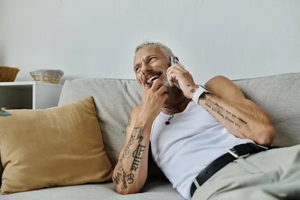 A mature gay man with tattoos and a grey beard sits on a couch at home, laughing as he talks on the phone. — Stock Photo