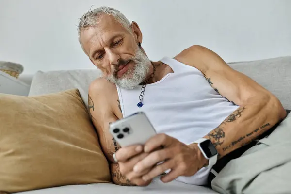 Um maduro tatuado gay homem com um cinza barba relaxa no um sofá em casa, usando seu telefone. — Fotografia de Stock