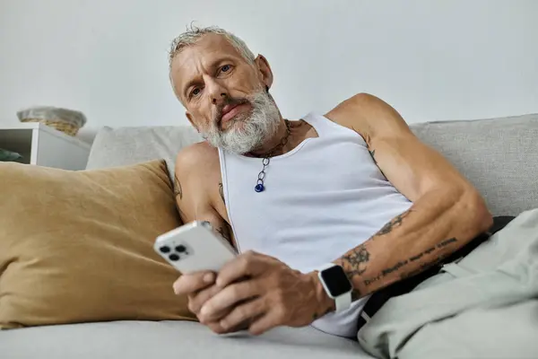 Um homem gay maduro com tatuagens e barba cinza relaxa em um sofá em casa, olhando para o telefone. — Fotografia de Stock
