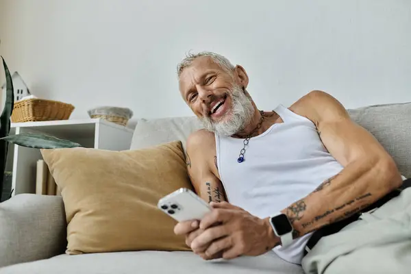 A mature gay man with tattoos and a grey beard relaxes on a couch at home, smiling and holding a smartphone. — Stock Photo