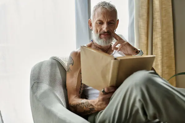 A mature gay man with tattoos and a grey beard relaxes on a couch at home, engrossed in a book. — Stock Photo