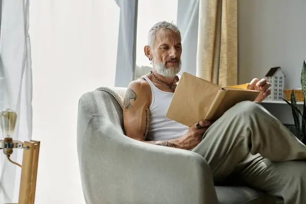 A mature gay man with tattoos and a grey beard relaxes on a couch at home, reading a book. — Stock Photo
