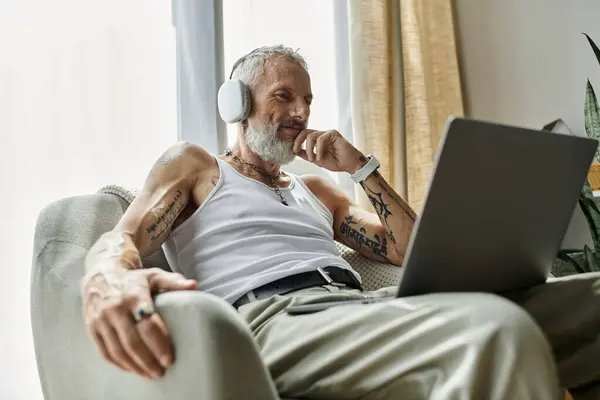Un homme gay mature avec des tatouages et une barbe grise se détend sur son canapé en utilisant un ordinateur portable. — Photo de stock