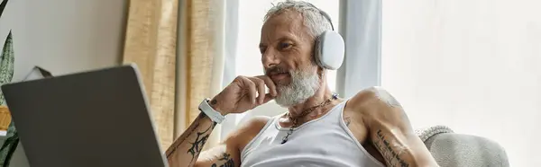 A mature gay man with tattoos and a grey beard relaxes on a couch at home, wearing headphones. — Stock Photo