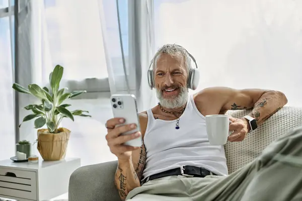 A tattooed gay man in a tank top smiles, listening to music on his phone with coffee on a couch at home. — Stock Photo