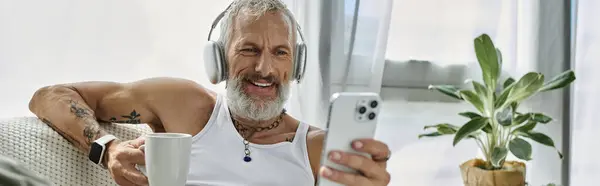 A mature man with grey beard and tattoos is smiling and relaxing on a couch at home. He is wearing headphones and looking at his phone. — Stock Photo