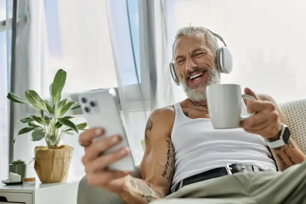 A mature gay man with tattoos and a grey beard relaxes on a couch at home, enjoying a cup of coffee and listening to music. — Stock Photo
