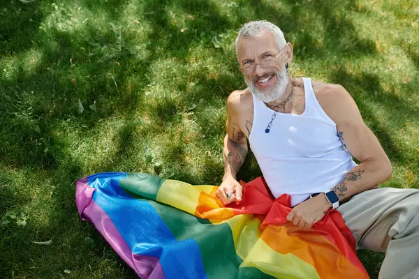 A mature gay man with tattoos and a grey beard is smiling and holding a rainbow flag outdoors in the grass. — Stock Photo