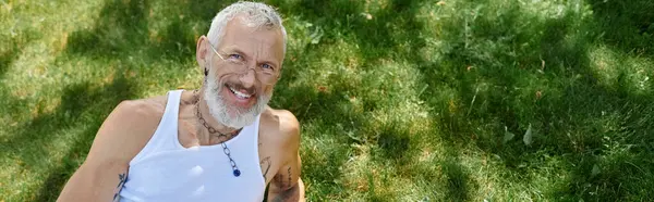 A mature gay man with tattoos and a grey beard smiles while sitting outdoors. — Stock Photo