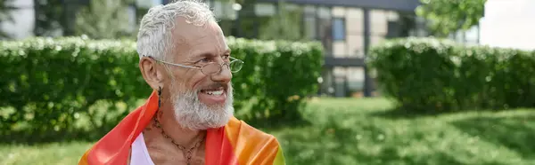 A mature gay man with tattoos and a grey beard smiles while holding a rainbow flag outdoors. — Stock Photo