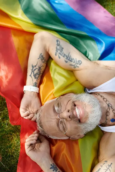A mature gay man with tattoos and a grey beard smiles while lying on a rainbow pride flag. — Stock Photo
