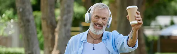 A mature man with tattoos and a grey beard smiling and holding a coffee cup. He is wearing headphones. — Stock Photo