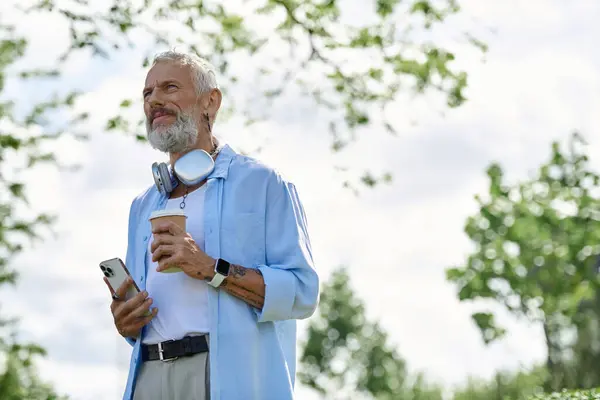 A mature gay man with tattoos and a grey beard sits on the grass outdoors, enjoying a cup of coffee. — Stock Photo