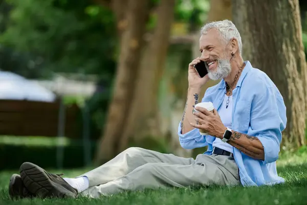 A mature gay man with tattoos and a grey beard sits on the grass, smiling while talking on his phone. — Stock Photo