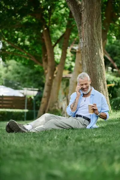 A mature gay man with tattoos and a grey beard sits on the grass outdoors. He is on a phone call and enjoying a coffee. — Stock Photo