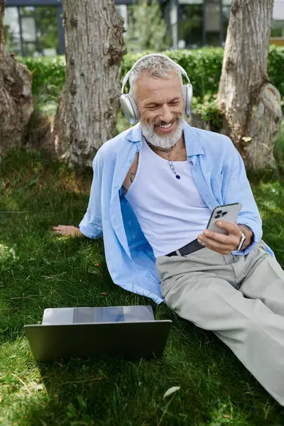 A mature gay man with tattoos and a grey beard sits on the grass, listening to music on headphones and using his smartphone. — Stock Photo