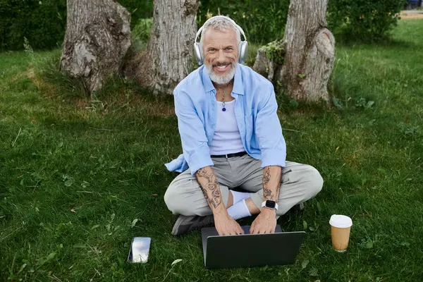 A mature gay man with tattoos and a gray beard sits on the grass in a park, working on his laptop while wearing headphones. — Stock Photo