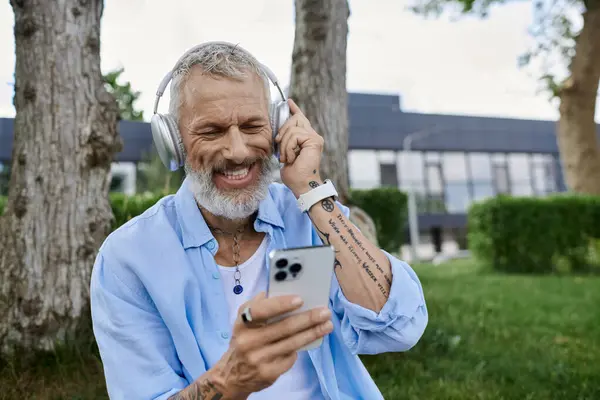 A man with tattoos and a grey beard sits on the grass, smiling while listening to music and looking at his phone. — Stock Photo