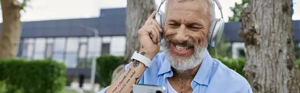 A mature man with grey beard and tattoos smiling brightly while listening to music through headphones. — Stock Photo