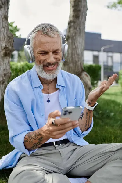 Um homem gay maduro com tatuagens e uma barba cinza senta-se na grama ao ar livre, usando fones de ouvido e sorrindo enquanto olha para o telefone. — Fotografia de Stock