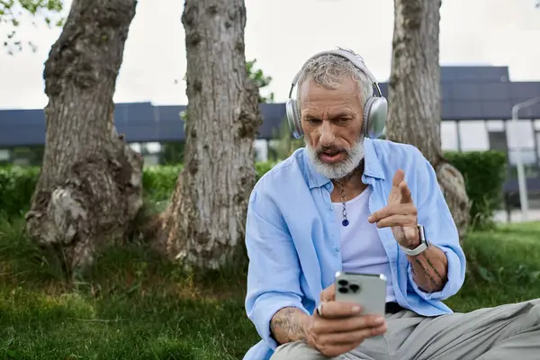 A mature man with tattoos and a grey beard sits on the grass in a park, wearing headphones and looking at his phone. — Stock Photo