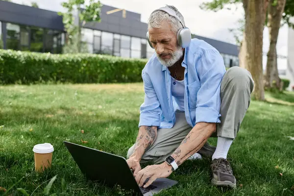 A mature man with tattoos and a grey beard sits on the grass outdoors, wearing headphones and typing on a laptop. — Stock Photo