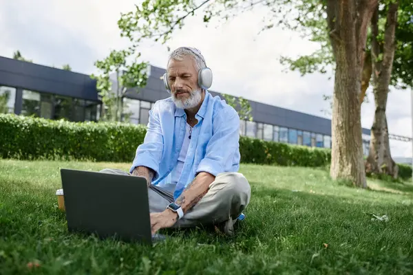 Un homme gay mature avec des tatouages et une barbe grise est assis sur l'herbe, travaillant sur son ordinateur portable. — Photo de stock