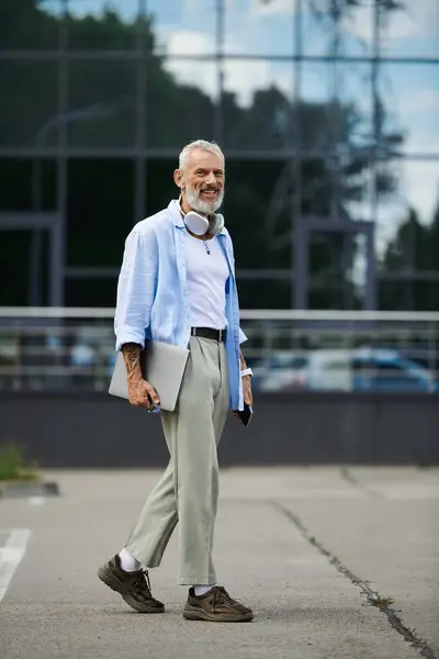 A mature gay man with a grey beard and tattoos walks outdoors with a laptop and headphones around his neck. — Stock Photo