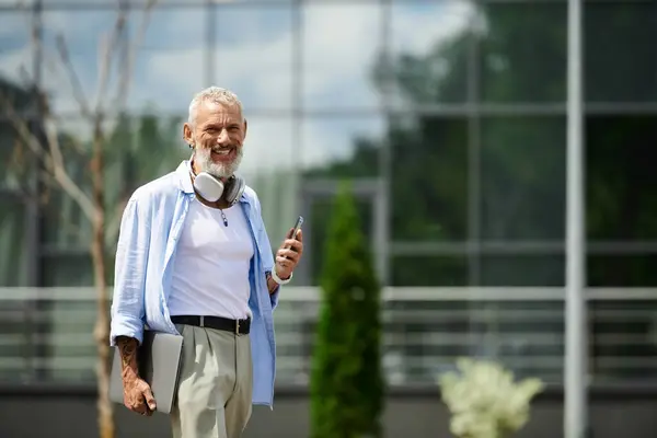 A mature man with tattoos and a grey beard smiles while holding a phone and a laptop. — Stock Photo