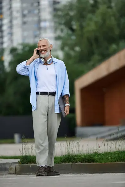 A tattooed, mature gay man with a grey beard smiles as he talks on his cell phone while walking outdoors in the city. — Stock Photo