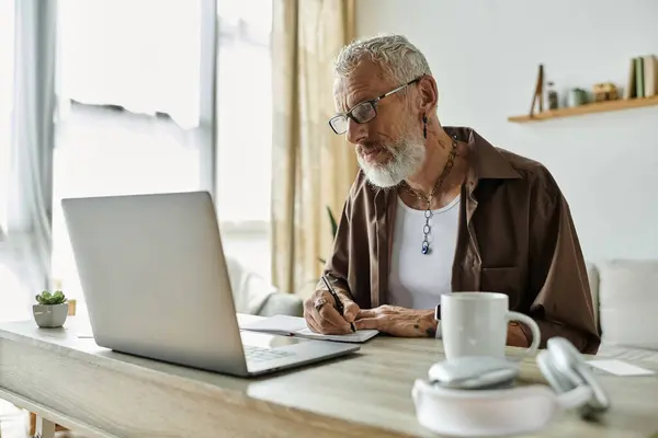 A mature gay man with tattoos and grey hair works remotely from home, using a laptop and taking notes. — Stock Photo