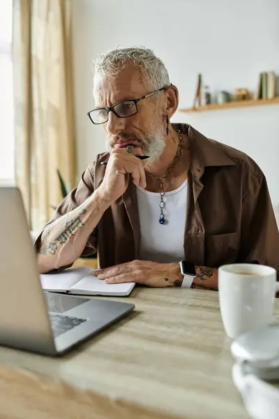 A mature gay man with gray hair and tattoos works remotely from home, focusing on his laptop and notebook. — Stock Photo