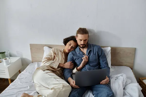 Couple relaxing in bed; woman resting on mans shoulder, he is using laptop. — Stock Photo