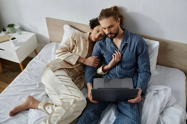 Ein Paar im Schlafanzug entspannt zusammen im Bett, wobei der Mann einen Laptop benutzt. — Stockfoto