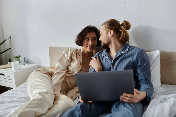 Una pareja cariñosa se relaja en la cama, acurrucándose y usando un portátil. - foto de stock