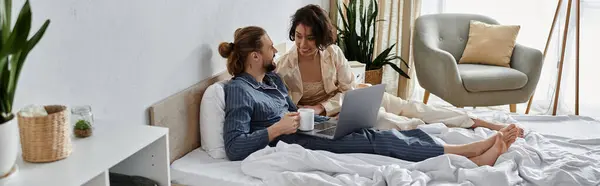 A couple in pajamas relaxes in bed, enjoying coffee and a laptop together. — Stock Photo