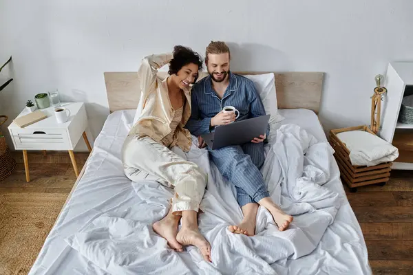 A loving couple cuddles in bed while enjoying coffee and using a laptop. — Stock Photo