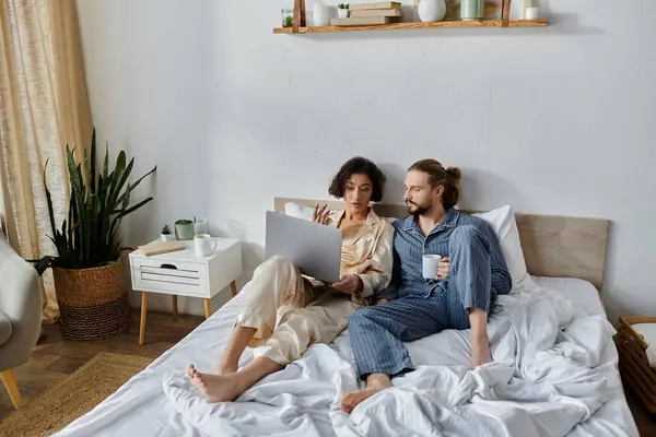 A loving couple relaxes in bed together, enjoying coffee and browsing a laptop. — Stock Photo