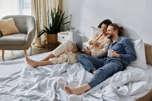 A couple relaxes in bed, watching something on a laptop. — Stock Photo