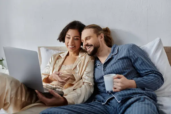 A loving couple in pajamas, relaxing in bed and enjoying coffee and a laptop. — Stock Photo