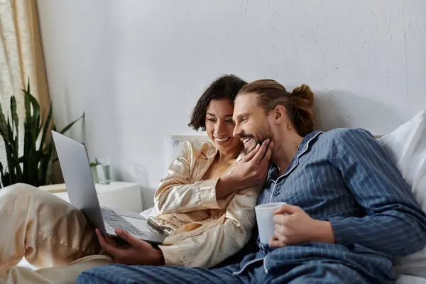 A couple cuddles in bed, enjoying a morning with coffee and a laptop. — Stock Photo
