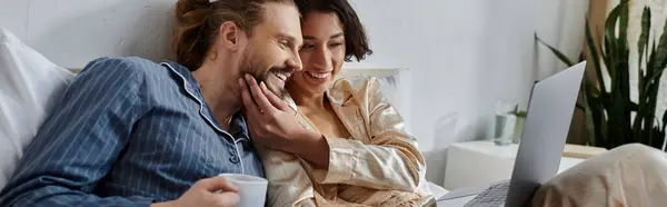 A loving couple relaxing in bed at home, with a laptop and a cup of coffee. — Stock Photo