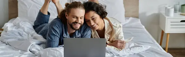 A couple relaxes in bed, cuddling and looking at a laptop. — Stock Photo