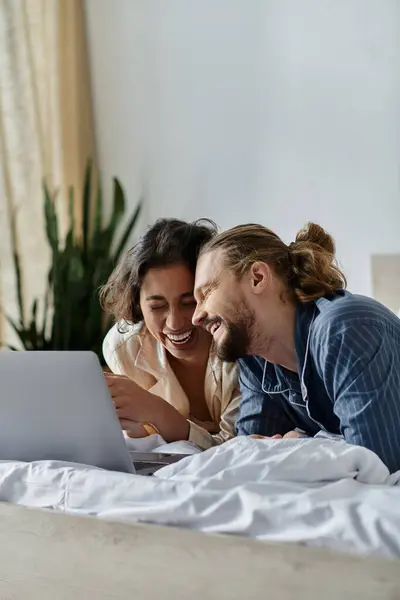 A loving couple relaxes in bed, laughing together while browsing the internet on a laptop. — Stock Photo