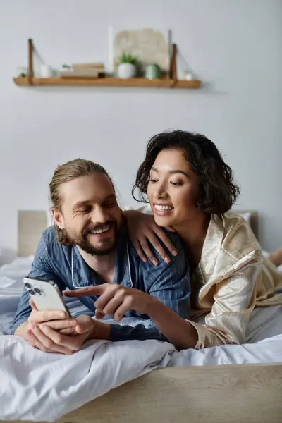 In bed, a couple relaxes: man on phone, woman smiling at him. — Stock Photo