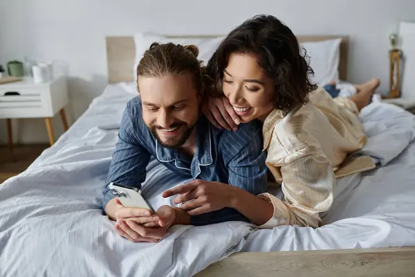 A couple in pajamas relaxes in bed, laughing and looking at a phone. — Stock Photo