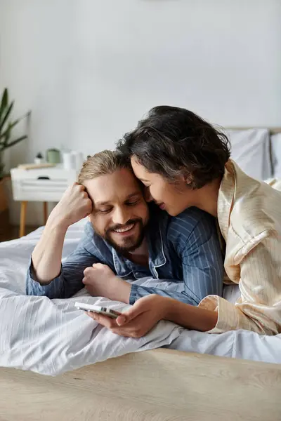 Un couple se détend au lit, la femme étreignant l'homme par derrière alors qu'il fait défiler au téléphone. — Photo de stock