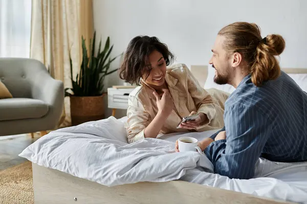 A couple cuddles up on a bed, enjoying a cup of coffee and a moment of laughter together. — Stock Photo